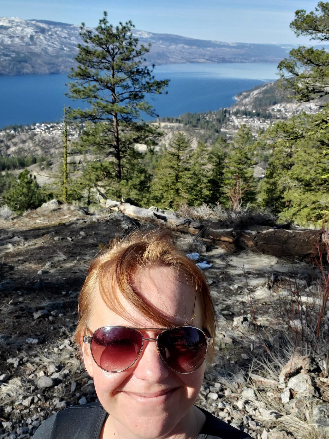 A smiling woman stands on a rocky mountain viewpoint overlooking a lake. 