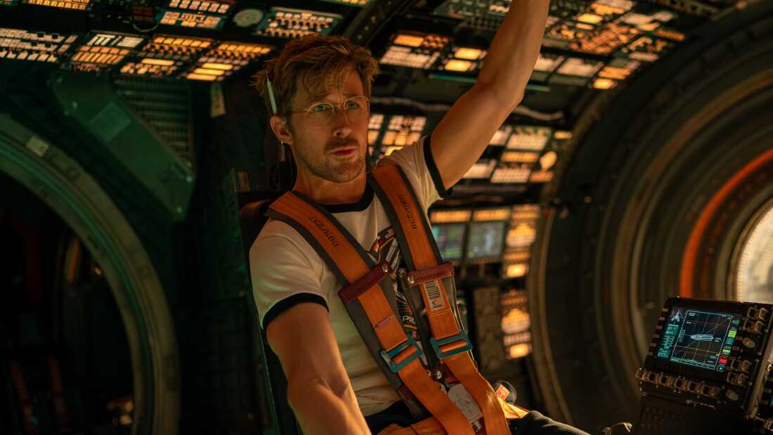 Ryan Gosling sits in the pilot seat of a dimly-lit spaceship. He is reaching above him with his left hand.