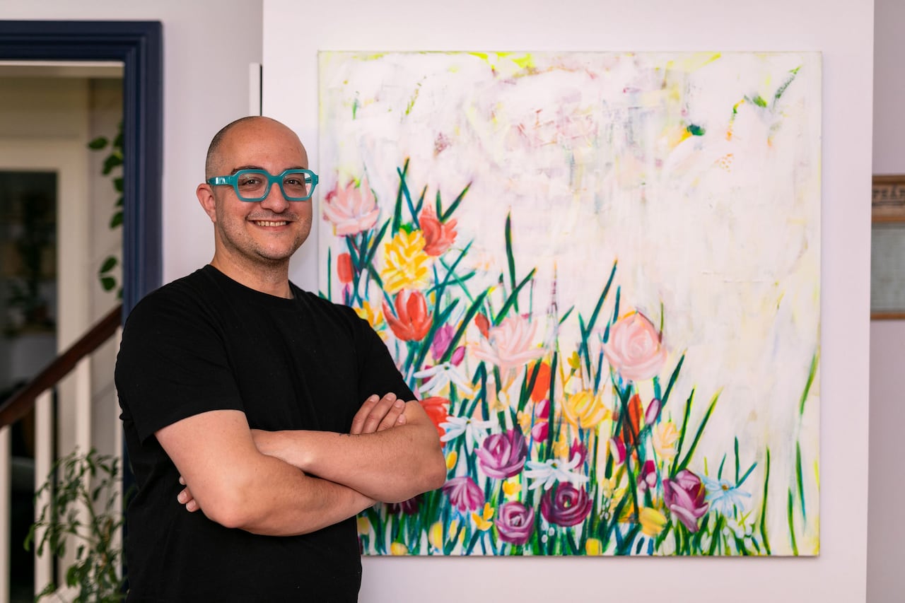 A man in a t-shirt and glasses stands beside a large canvas painted with flowers.
