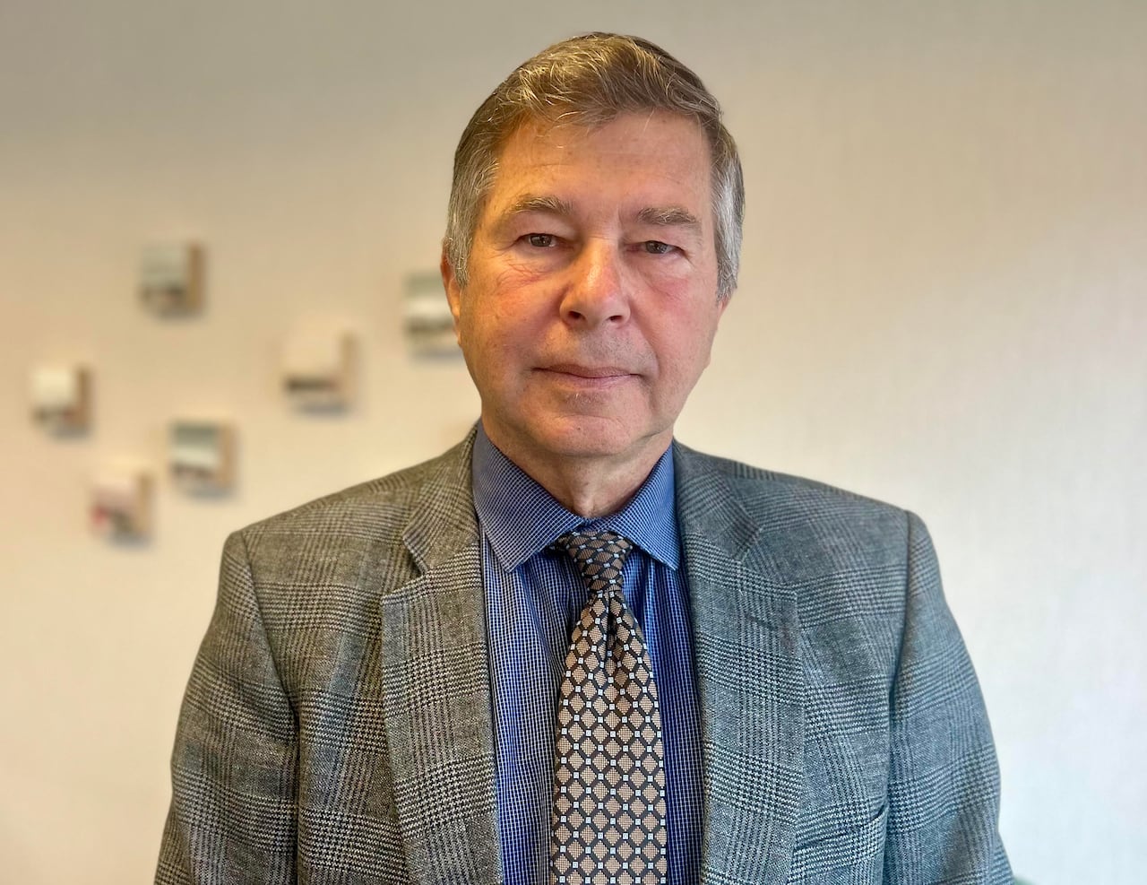 A man with grey hair, and a suit and tie, looks at the camera with a serious face, standing inside an office.  
