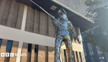 A metal statue showing a life-sized man dressed in steelworkers' uniform and holding a long pole with both hands. There is a light-coloured brick building in the background.