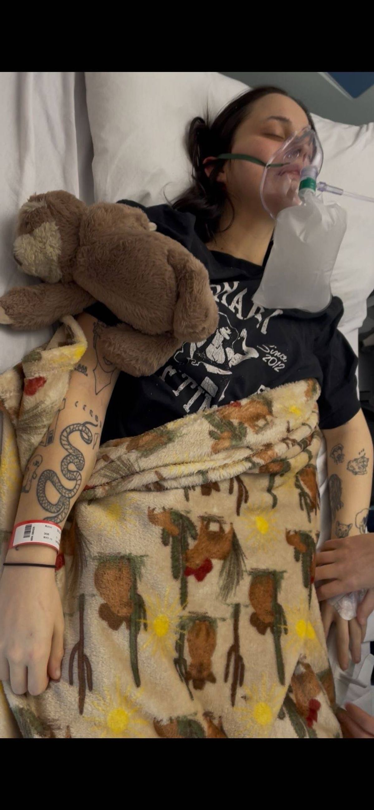 An individual lying in a hospital bed holding a brown plush toy. The person is wearing a black t-shirt with a graphic design. Nearby, a clear plastic container is visible.