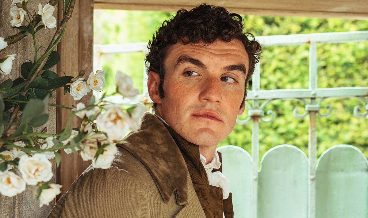 An individual with curly hair is standing and gazing towards the left, perhaps inside a room. The person is dressed in a brown jacket with a white inner shirt. In the background, there is a white-framed window adorned with white flowers, and lush greenery is visible beyond the window.