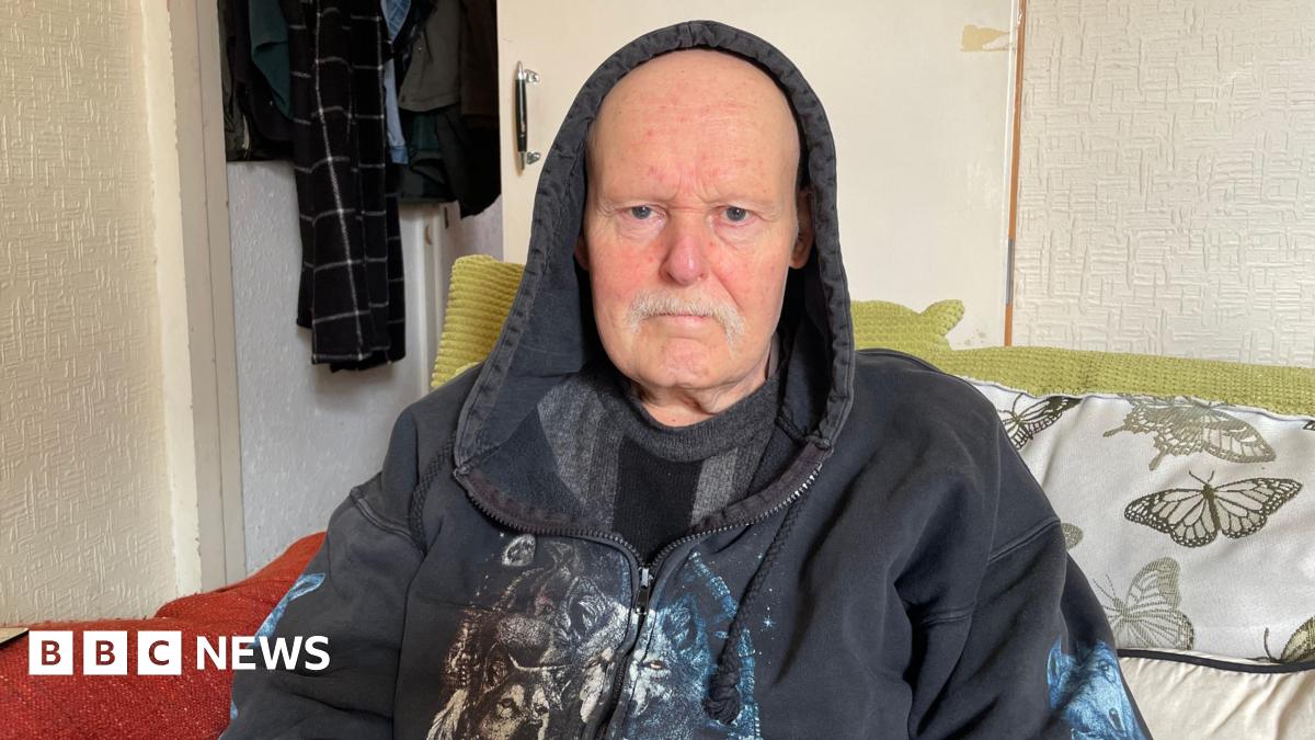 Tunde Olawoye stood behind the counter at his pharmacy wearing a brown fleece jumper. There are shelves of drugs and medicine behind him.