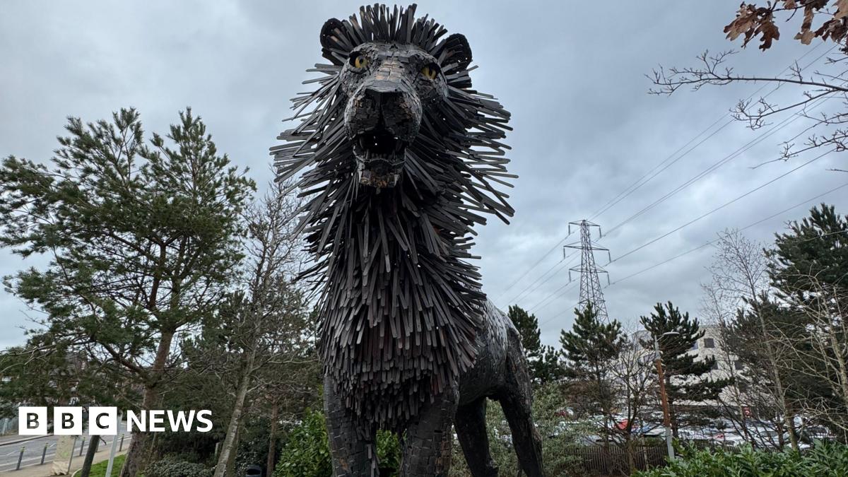 A statue of the Gruffalo , a mythical creature with a large head, orange eyes, a wide month, a round torso, horns, tusks and large claws.  The statue is standing in a wooded area with one paw leaning on a wooden post.