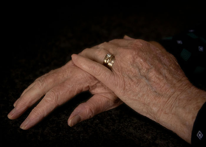 A close-up of elderly hands resting on a dark surface, highlighting signs of aging and medical secrets.