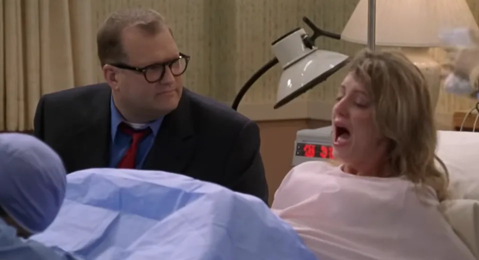 A man in glasses and a suit sits beside a woman in a hospital bed who appears to be in labor, with medical staff attending