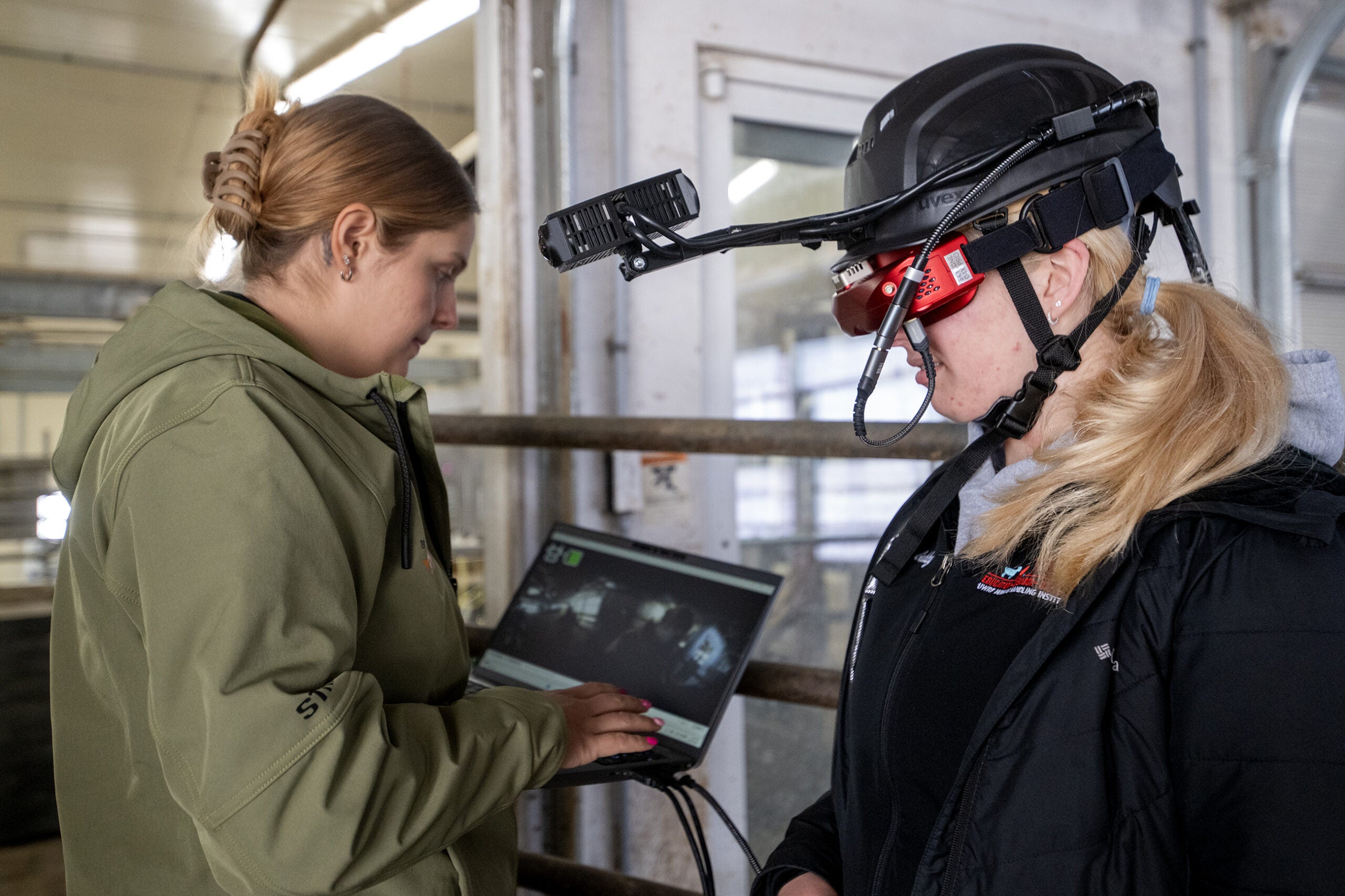 Two women stand indoors; one operates a laptop while the other wears a black helmet with a mounted camera and display.