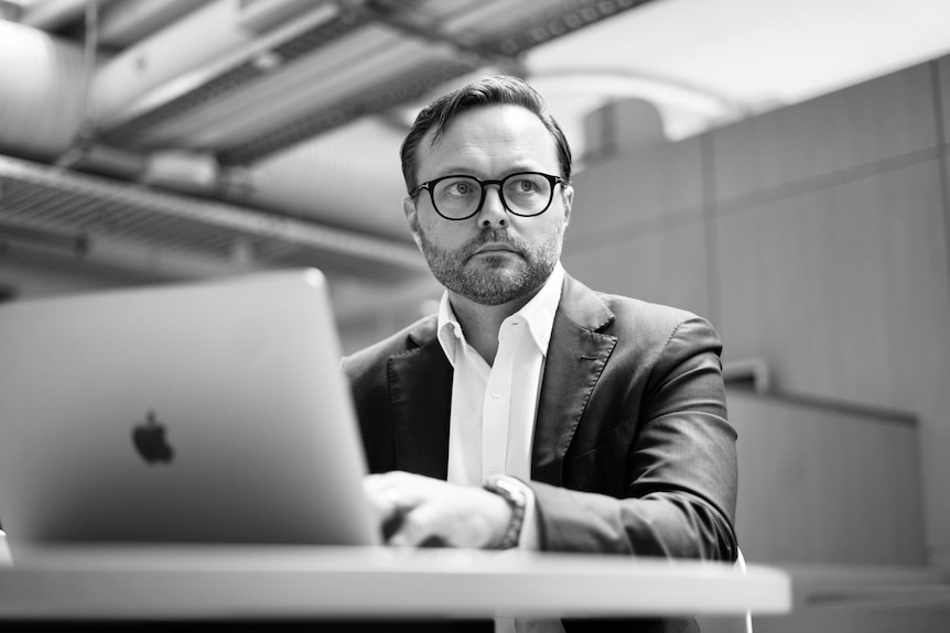 A black-and-white shot of a dark-haired man in glasses and a suit sitting at a laptop in an office.