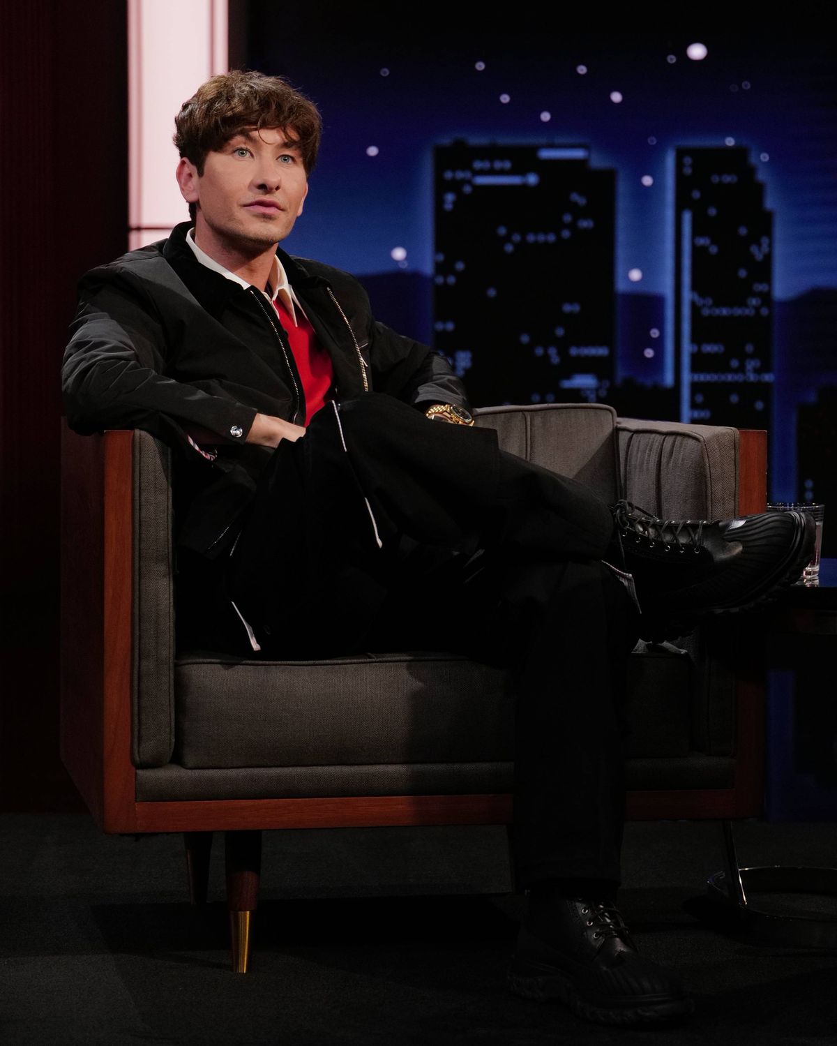 An individual dressed in a formal suit, seated on a chair with one leg crossed over the other, against a backdrop featuring a cityscape at night.