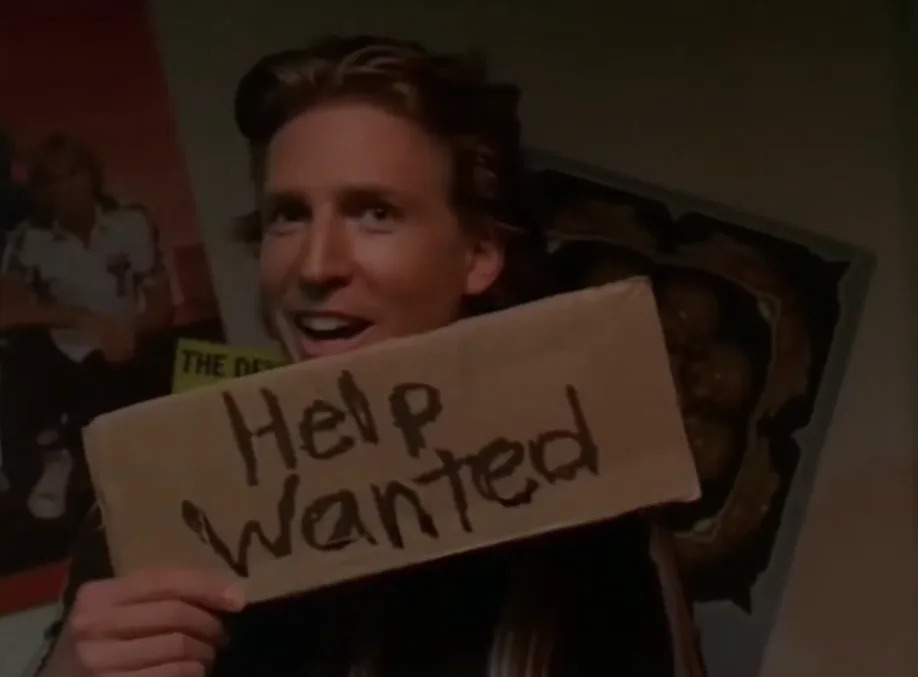 Person holding a "Help Wanted" sign, smiling, with posters visible in the background