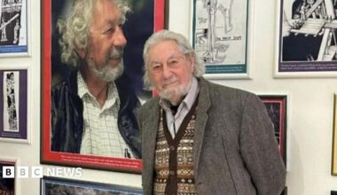 Curator Matthew Richardson, a man with grey hair, on the left and Major Shane Lucas, a man with long grey hair and a beard, on the right stand in front of a wall of paintings.