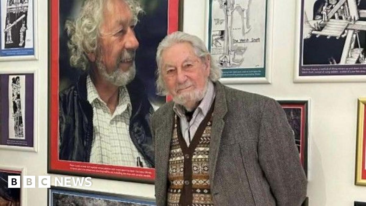 Curator Matthew Richardson, a man with grey hair, on the left and Major Shane Lucas, a man with long grey hair and a beard, on the right stand in front of a wall of paintings.