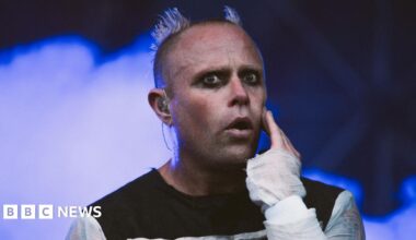 Keith Flint is on stage while wearing a black top with white sleeves. He has dark eyeliner on and his head is shaved, except for two parts which stand up like devil horns. Blue smoke billows behind him.