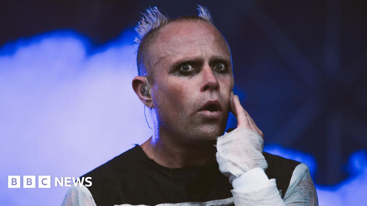Keith Flint is on stage while wearing a black top with white sleeves. He has dark eyeliner on and his head is shaved, except for two parts which stand up like devil horns. Blue smoke billows behind him.