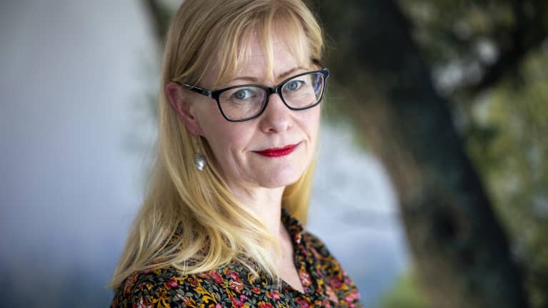 Woman with blonde hair and eyeglasses wearing a flower-patterned shirt looking into the camera.