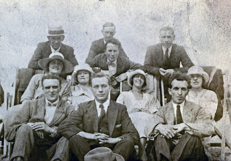 A vintage black-and-white photo of eleven men and women in formal early 20th-century attire, seated and standing in rows, posing for a group portrait outdoors.