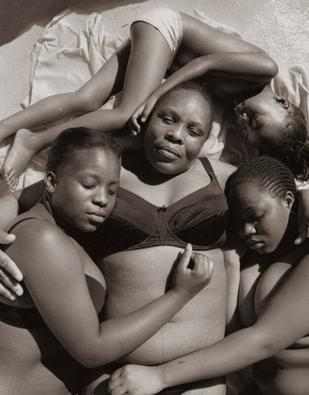A black woman lies on her back with her eyes closed, surrounded closely by three children, all embracing her and each other, conveying warmth and intimacy. The image is in black and white.