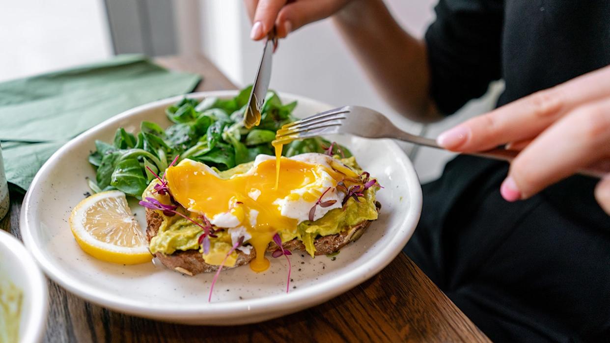 Avocado toast topped with poached eggs and runny yolk served with fresh greens and lemon slice on a white plate.