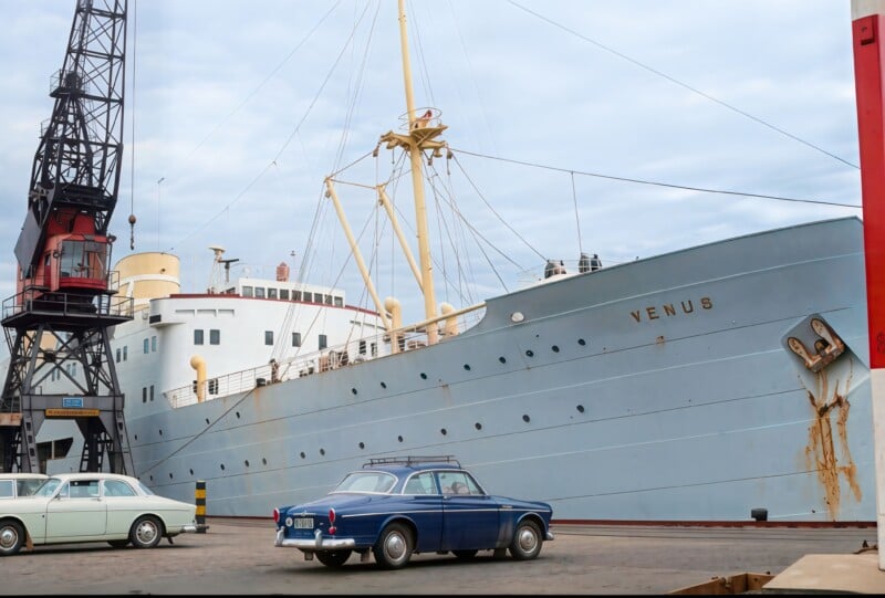 A large ship named VENUS docked at a port, with two vintage cars—a blue one in the foreground and a white one beside it—parked nearby. A crane stands to the left, and the sky is overcast.