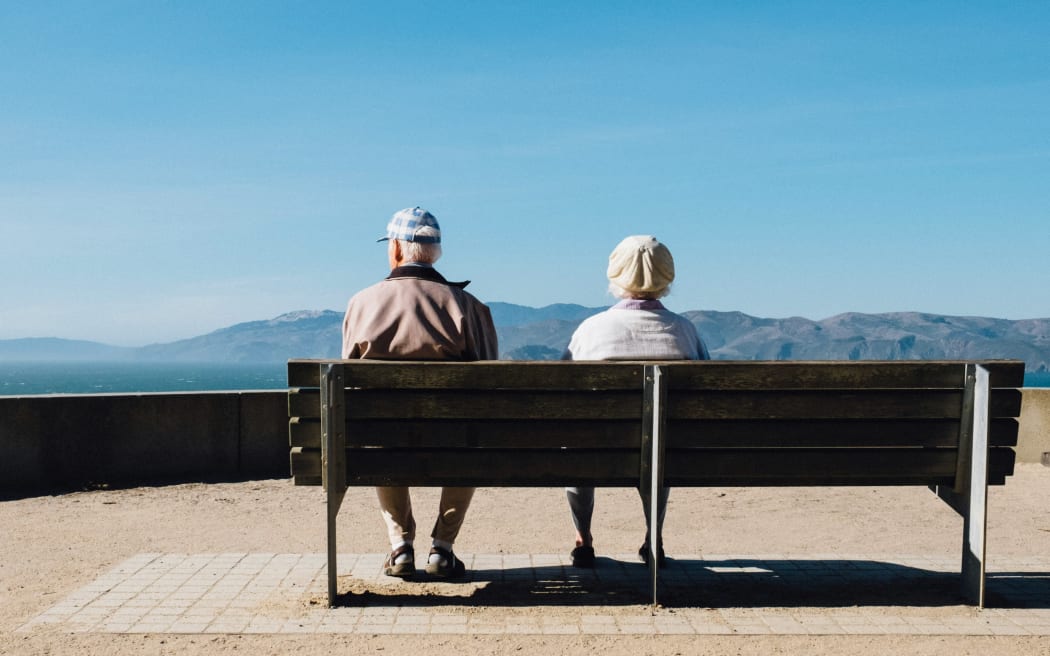 Two elderly people sit on a bench looking at a view of sea and hills.