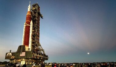a rocket stands on a launch tower against a fading blue sky and a distant full moon casting rays outward.