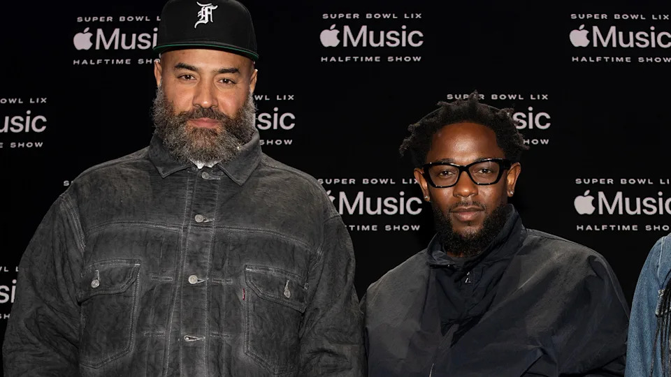 Ebro and Kendrick Lamar stand in front of an Apple Music Super Bowl Halftime Show backdrop. Both are wearing dark jackets and one has glasses.