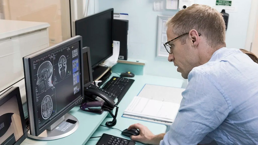 a light-skinned man sits at a computer screen showing a human head and brain