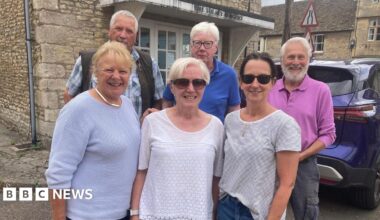 A group of people - three women and three men - stand outside an cold Cotswold stone building with a car outside and a sign on it that says 'the Tolsey Surgery'