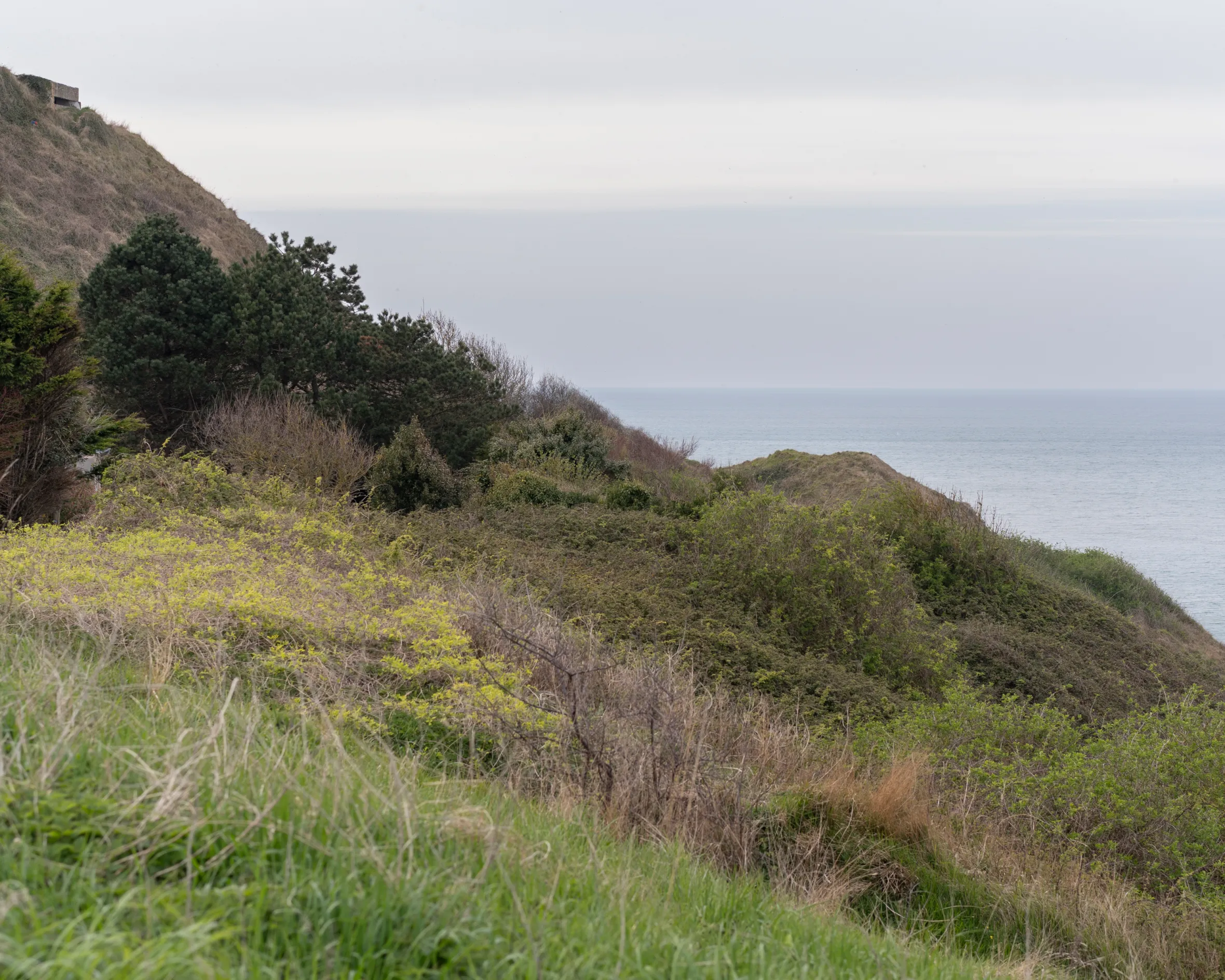 A grassy hill, topped by a concrete bunker, overlooks the calm sea at Port-en-Bessin, France.