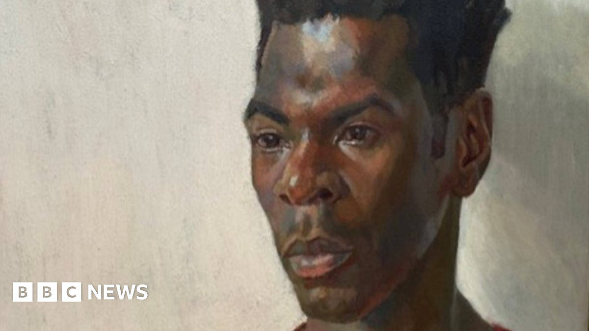 A head and shoulders portrait of the artist Nahem Shoa shows him wearing a plain red top, looking slightly off to the right, his gaze fixed on somthing or somone we can't see.