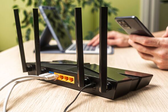 Wireless internet router on light office desk, user in background works on laptop and phone, configuring stable home network con