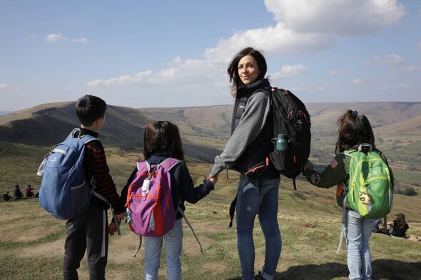 Alt text: A woman and three children, all equipped with backpacks, are standing on a grassy hill under a blue sky, with distant 