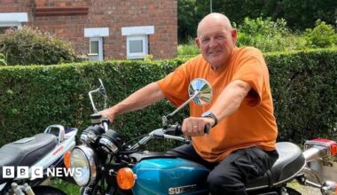 Tom Frith smiles as he poses on an old motorcycle in front of a hedge outside a garden.