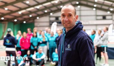 Tim Howe standing in front of a group of people in a sports hall. He smiles at the camera and wears a dark-blue hoodie which has The Longevity Games logo on it.