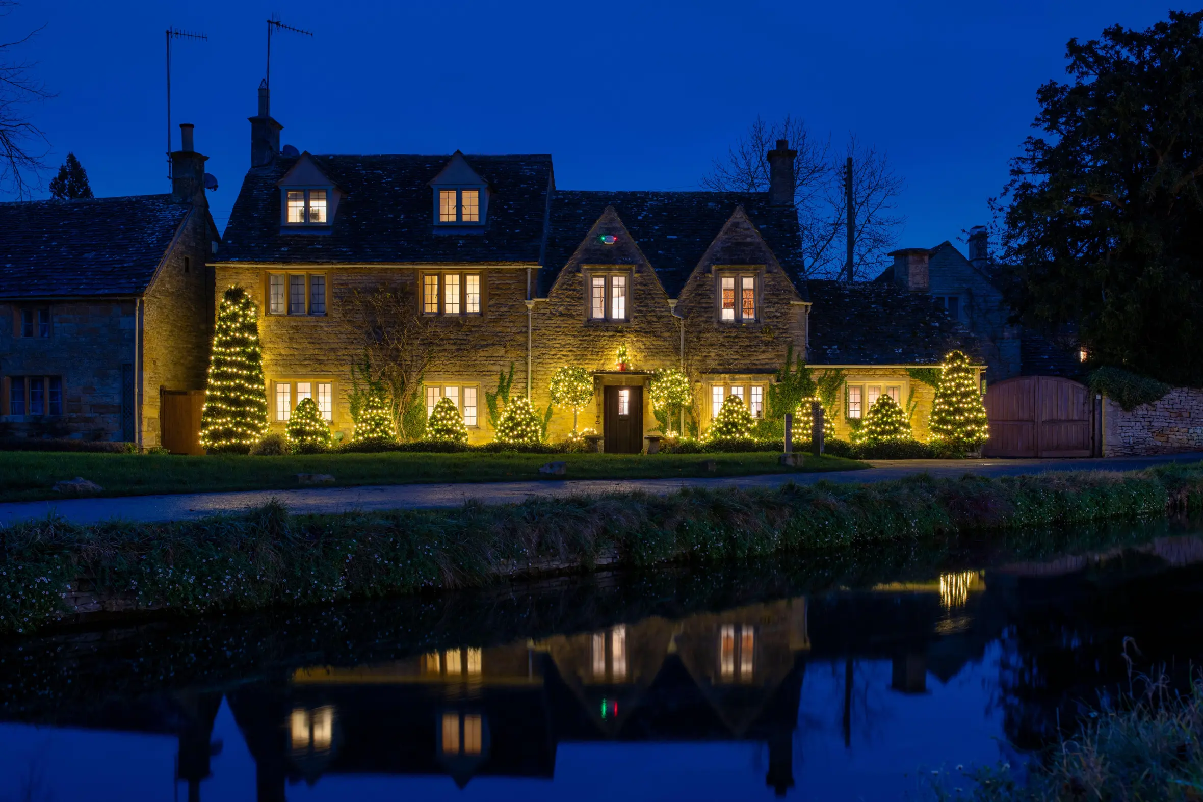Cotswold cottage in Lower Slaughter decorated with Christmas lights at dusk, reflected in the water.