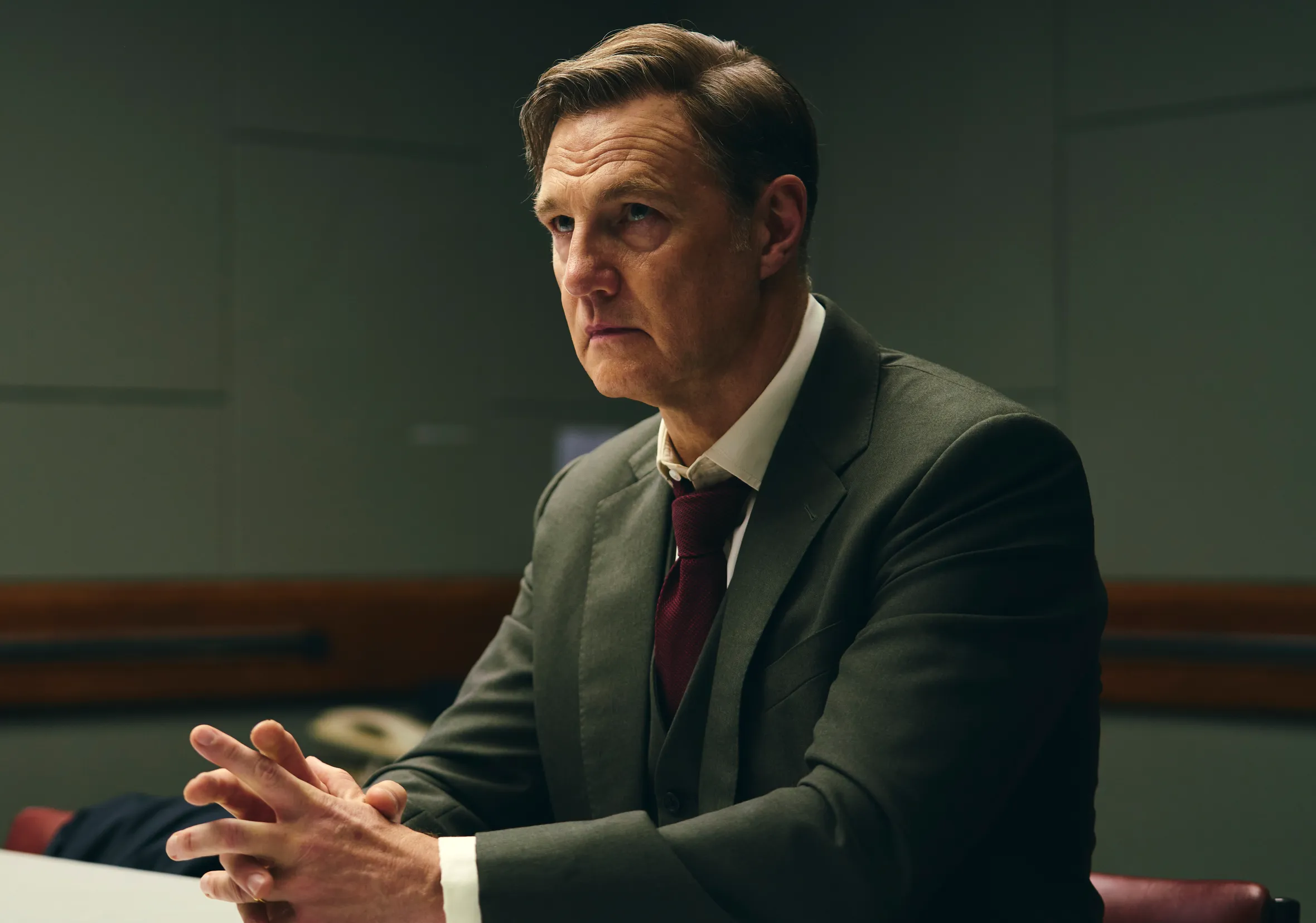 David Morrissey as Michael Polly, wearing a suit, seated at a desk with his hands clasped.