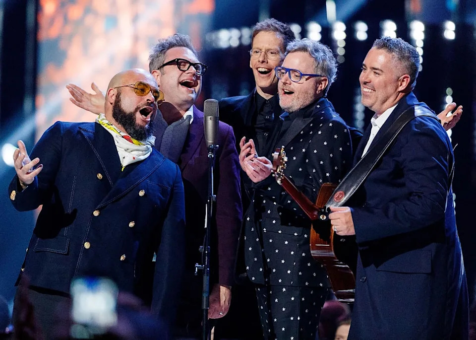 Group of musicians performing on stage, wearing suits and smiling, with one holding a guitar