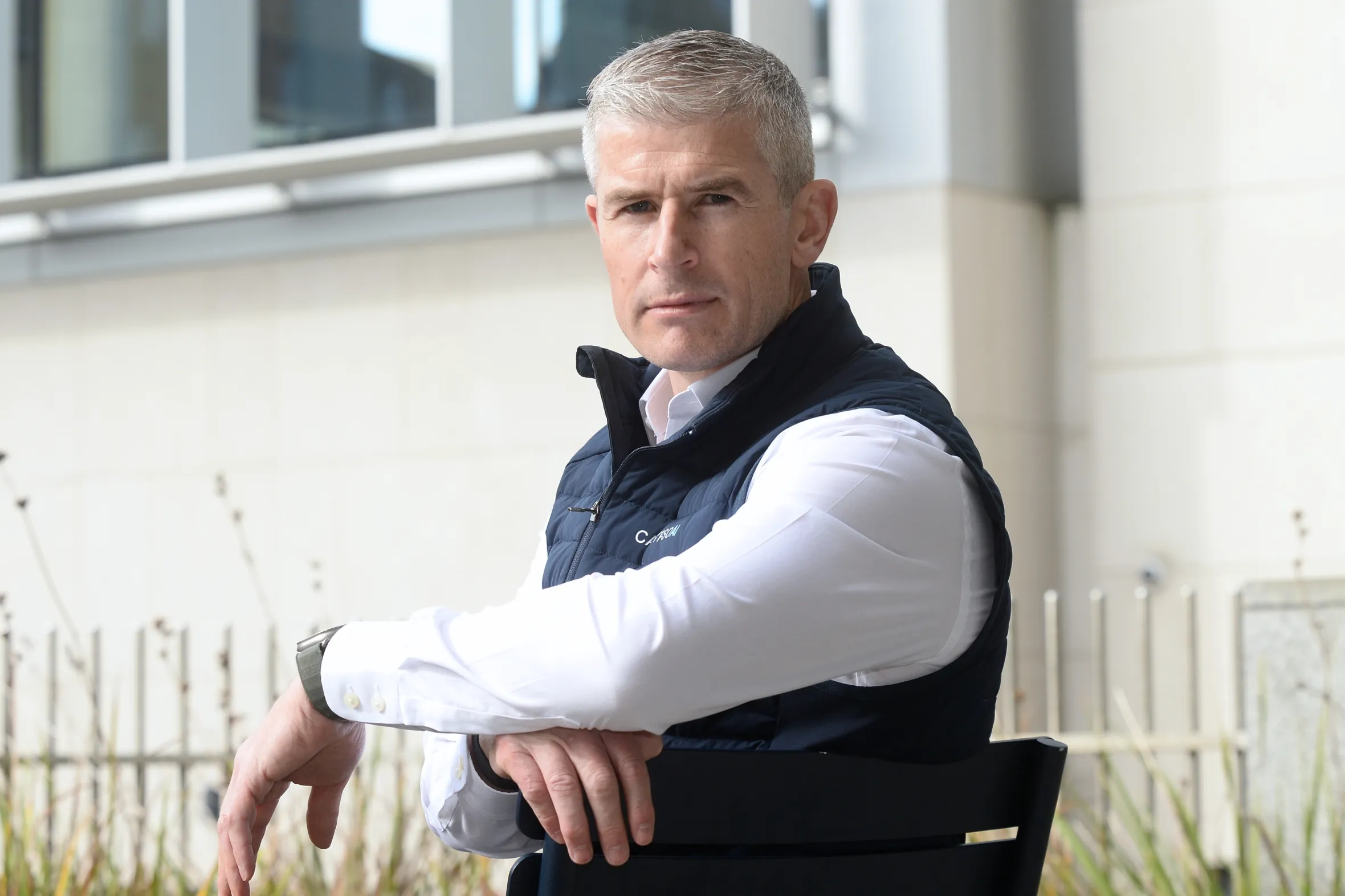 Donnchadh Casey, CEO of CalypsoAI, sits wearing a white shirt and navy vest with his arms resting on the back of a chair.