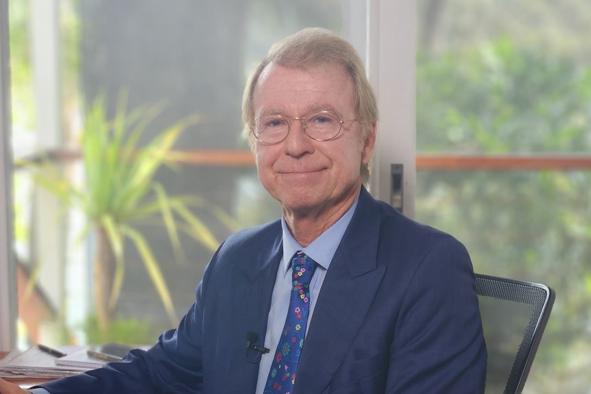 A middle-aged, sandy-haired man in glasses and a suit sits in an office.