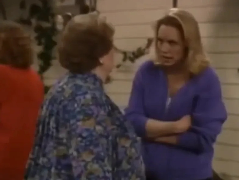 Two people are engaged in a conversation on a sitcom set. One person is wearing a patterned blouse, and the other is wearing a casual zip-up jacket