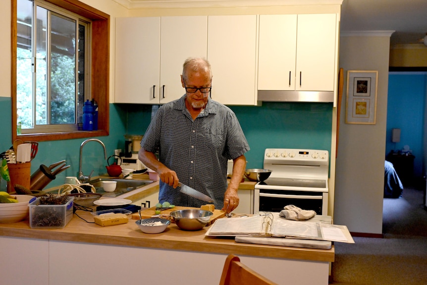 Geoff stands in the kitchen cutting vegetables in a dated kitchen, white cabinets, old stove, turquoise blue splashback.