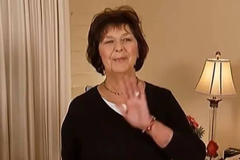 An elderly woman with short hair, dressed in a black top, stands indoors and gestures with her hand while smiling. A lamp with a white shade is visible on her right side, and a curtain is positioned to her left.