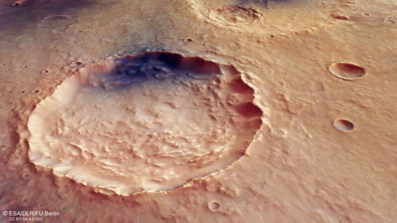 A high-resolution image shows a large, round impact crater on a rocky, reddish-brown Martian surface with several smaller craters nearby, under soft lighting.