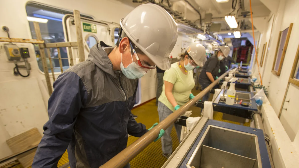 Above left, Oscar Cavazos (Marine Laboratory Specialist, IODP JRSO) joins other marine techs in preparing the core new to be sectioned on the catwalk. Photo Credit: Erick Bravo, IODP JRSO