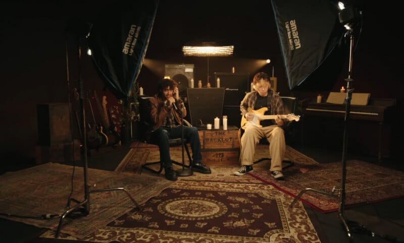Two musicians sit on patterned rugs in a dimly lit studio, surrounded by guitars, amplifiers, candles, and a piano. Studio lighting and equipment are visible, creating a cozy, creative atmosphere.