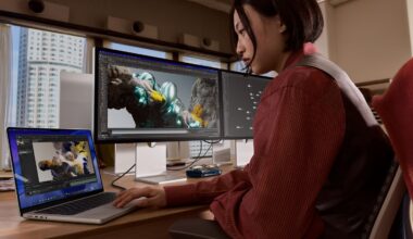 A person sits at a desk using a laptop and two large monitors, working on 3D digital artwork with colorful graphics displayed on the screens in an office with large windows and city views.