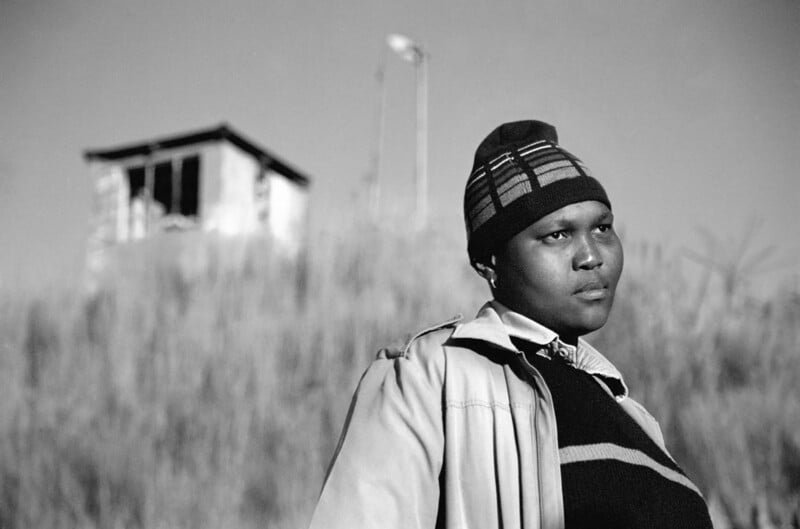 A person wearing a striped beanie, jacket, and sweater stands outdoors with tall grass and a small, weathered building in the background. The image is in black and white.