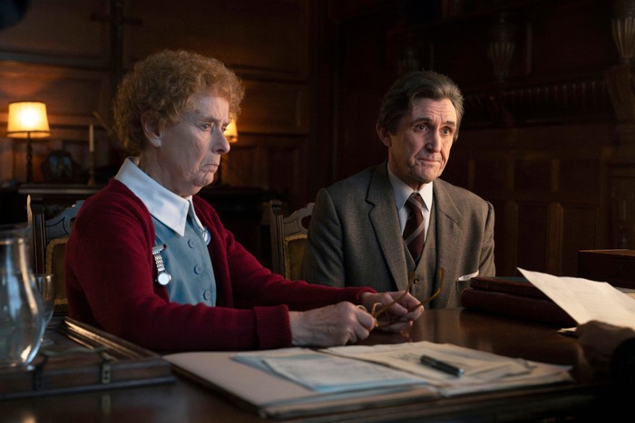 Two people sit at a wooden table reviewing paperwork in a warmly lit room, looking serious as they listen to someone off-camera.