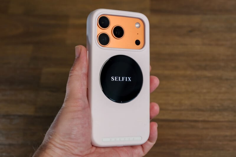 A hand holds a smartphone with a peach-colored back, three cameras, and a white case. A black circular device labeled "SELFIX" is attached to the back. The background is a wooden surface.